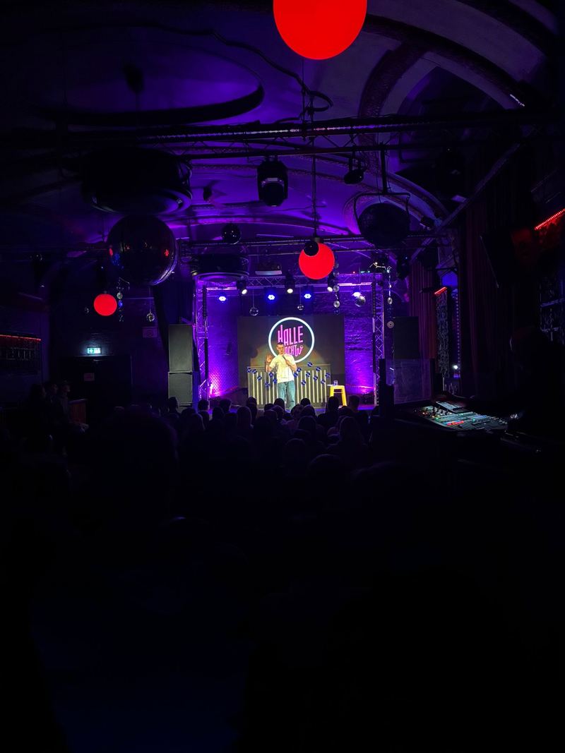 Comedian mit Mikrofon auf kleiner Bühne, Neonschriftzug „Halle lacht